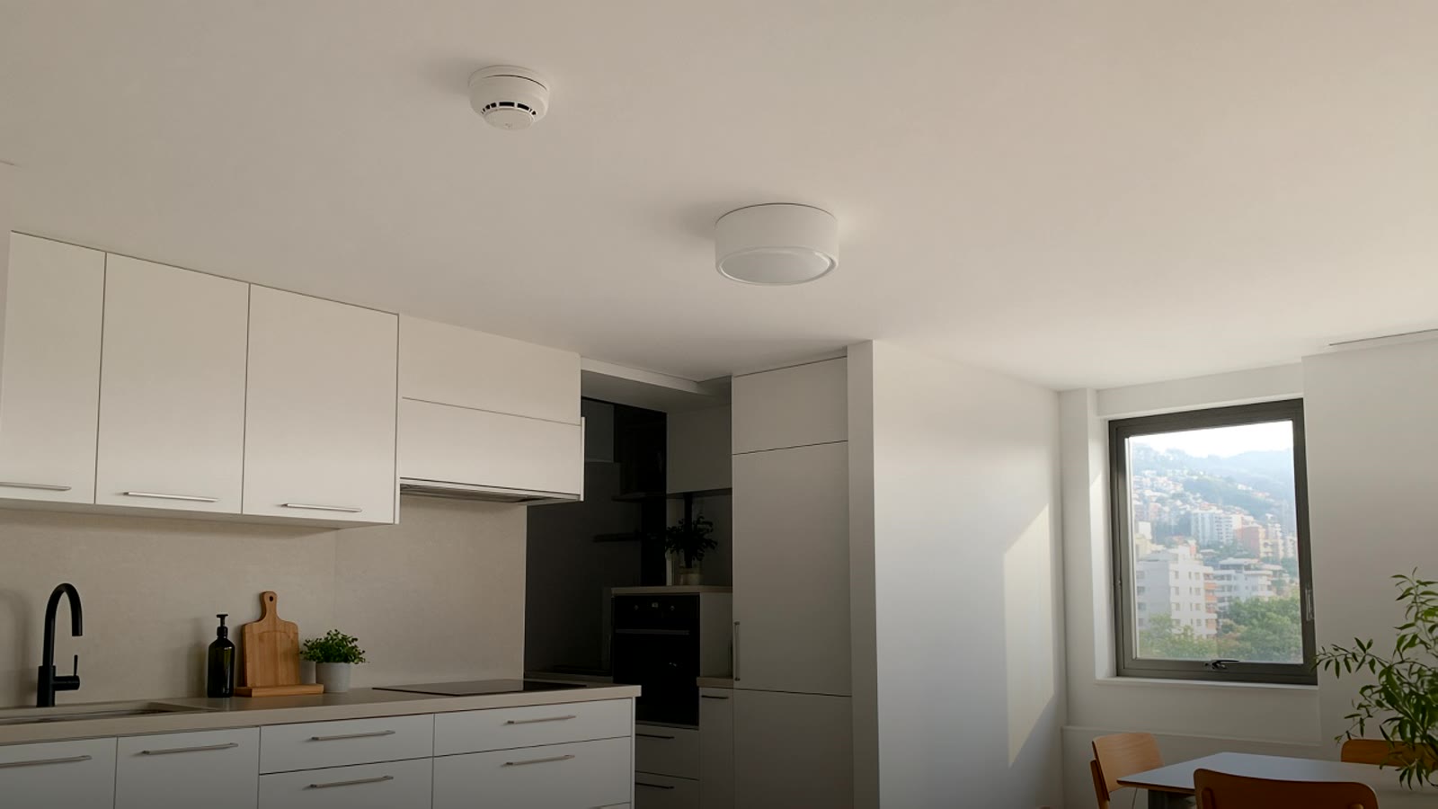 White ceiling of an open kitchen in an apartment, with a small round smoke detector near the light fixture and afternoon light coming through the window